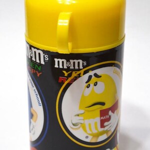 May include: A cylindrical M&M's container with a yellow lid and handle. The container is black with M&M's branding and graphics of the candy characters. The text "m&m's" and "YET READY" are visible.