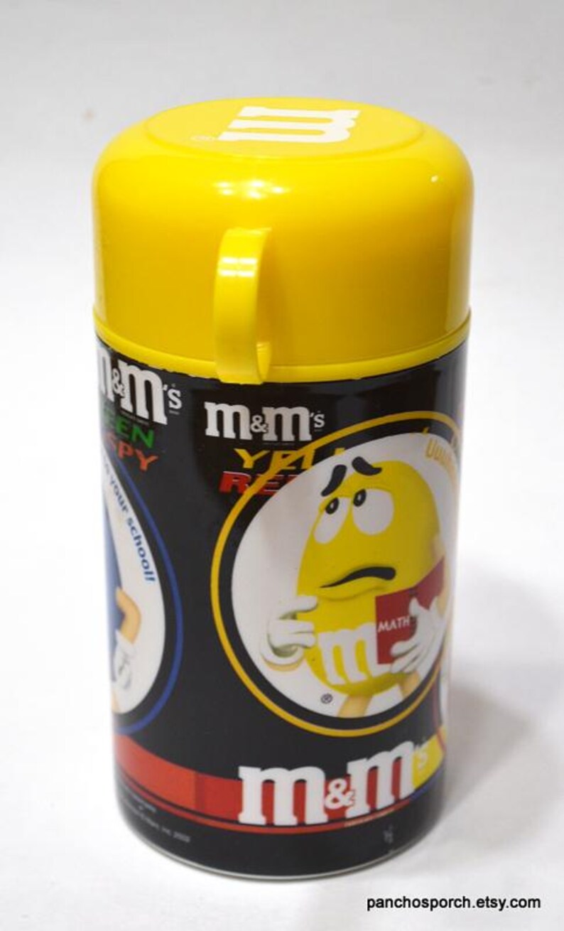 May include: A cylindrical M&M's container with a yellow lid and handle. The container is black with M&M's branding and graphics of the candy characters. The text "m&m's" and "YET READY" are visible.