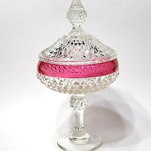 Peut inclure: Une compote en verre transparent avec un motif taillé en diamant et une bande rose vif. La compote est dotée d'un pied et d'un couvercle avec un bouton pointu, idéale pour servir des desserts ou exposer des objets décoratifs.