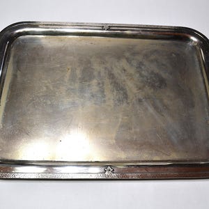 May include: A rectangular silver serving tray with two curved handles. The tray has a slightly raised edge and a decorative border. The surface of the tray is reflective, showing signs of wear. A classic piece for serving food and drinks.