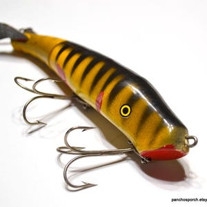 May include: A vintage fishing lure with a yellow and black striped body, red accents, and three treble hooks. The lure has a black head and a small propeller at the end. The eye is yellow with a black center.