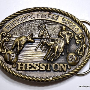May include: An oval brass-toned belt buckle featuring a rope-style border. The center depicts a rodeo scene with a rider on horseback roping a steer. Text reads NATIONAL FINALS RODEO, 19, 80, and HESSTON. The metal has a raised, textured relief.
