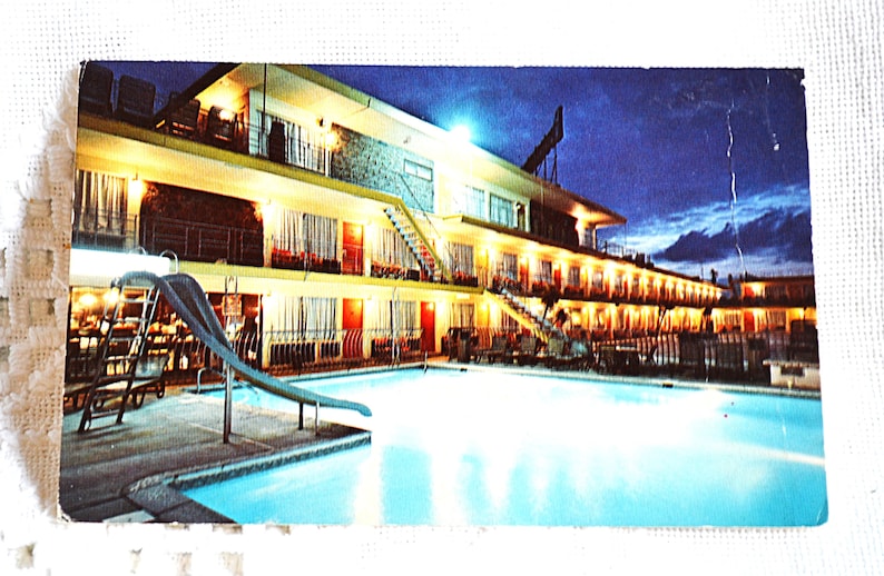 May include: Vintage postcard featuring a motel at night. The multi-story building has lit rooms, a swimming pool, and a water slide. The sky is dark blue, and the building has a retro aesthetic.