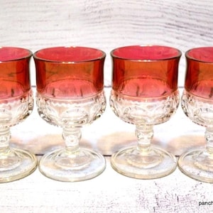 May include: Four vintage wine glasses with ruby red bowls and clear, patterned bases. The glasses have a classic design, perfect for serving wine or other beverages. The stemware is arranged on a white wooden surface.