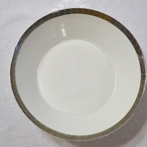 May include: A white ceramic bowl with a silver rim featuring a repeating leaf pattern. The bowl has a wide, shallow design, suitable for serving soups or salads. The rim adds a touch of elegance to the tableware.