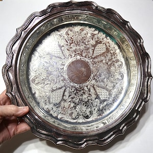 May include: Silver-plated serving tray with an ornate, engraved floral and scroll design. The tray has a scalloped edge and a central, circular area. The tray is held by a hand.