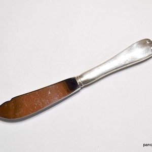 May include: A silver butter knife with a rounded handle and a wide, slightly curved blade. The knife is made of a shiny metal and is set against a white background. The handle has a decorative design.