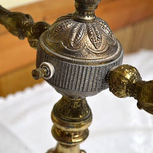 Puede incluir: Una base de l&aacute;mpara de lat&oacute;n con un eje central ornamentado que presenta una c&uacute;pula con patr&oacute;n de hojas, una secci&oacute;n cil&iacute;ndrica estriada y un peque&ntilde;o interruptor de bot&oacute;n plateado. La estructura incluye varios brazos con detalles.
