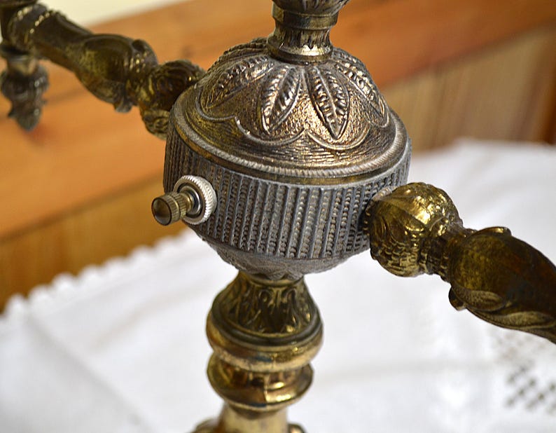 Puede incluir: Una base de l&aacute;mpara de lat&oacute;n con un eje central ornamentado que presenta una c&uacute;pula con patr&oacute;n de hojas, una secci&oacute;n cil&iacute;ndrica estriada y un peque&ntilde;o interruptor de bot&oacute;n plateado. La estructura incluye varios brazos con detalles.
