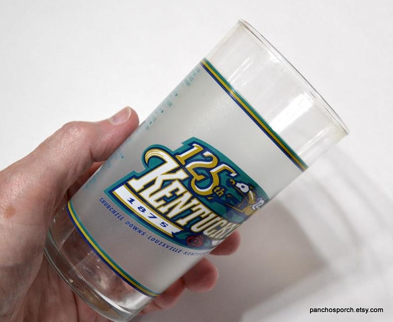 Pu&ograve; includere: Un bicchiere di vetro trasparente con una fascia bianca smerigliata con la scritta "125 Kentucky" in verde acqua e oro, con "Churchill Downs Louisville Kentucky 1875" sotto. Il bicchiere ha un bordo giallo e verde.