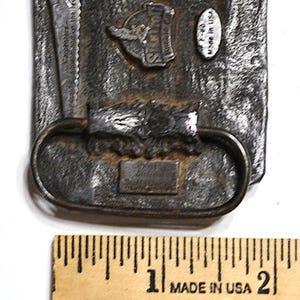May include: A rectangular, textured metal belt buckle with a saw and a logo that says "Sperry". The buckle has an oval loop and a small rectangular plate. The text "Made in USA" is visible. A wooden ruler is in the foreground.