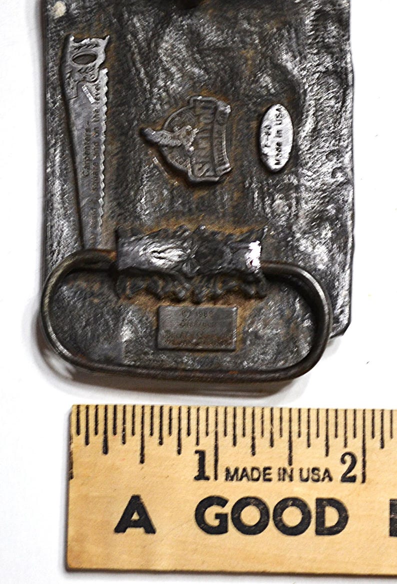 May include: A rectangular, textured metal belt buckle with a saw and a logo that says "Sperry". The buckle has an oval loop and a small rectangular plate. The text "Made in USA" is visible. A wooden ruler is in the foreground.