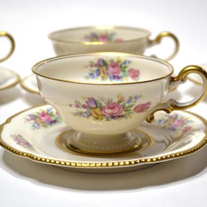 Könnte beinhalten: Ein Set aus cremefarbenen Porzellantassen und Untertassen mit einem zarten Blumenmuster aus rosa Rosen, gelben Blüten und blauen Akzenten. Jedes Teil ist mit einem goldfarbenen Rand und einem gold bemalten Henkel an den Tassen versehen.