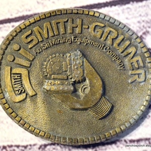 May include: A round, brass-toned metal medallion featuring the embossed text SMITH-GRUNER and An Sii Mining Equipment Company. The center displays a raised graphic of a mining drill bit, with the word SMITH integrated into the design.