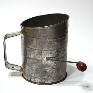 May include: A vintage metal flour sifter with a cylindrical body, a side handle, and a rotating crank featuring a dark wooden knob. The exterior surface shows signs of wear and oxidation, with embossed measurement markings for one, two, and three cups.
