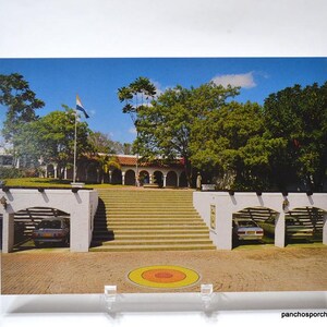 May include: A vintage postcard featuring a stately building with a grand staircase and arched entrance. Two cars are parked under white archways. A flag waves in the breeze against a backdrop of lush green trees and a clear blue sky.