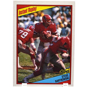 May include: A Topps trading card featuring a football player in a red uniform, holding a football. The card has a red and yellow border with the words "Instant Replay" at the top. The player is Steve Bartkowski.