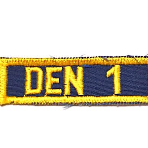 May include: A blue and yellow embroidered patch with the text "DEN 1" on it.