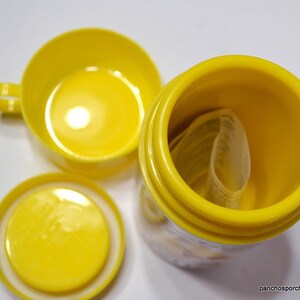 May include: Bright yellow ceramic cup with a handle, a matching lid, and a cylindrical container. The container is open, revealing a rolled-up paper inside. The items are arranged on a white surface.