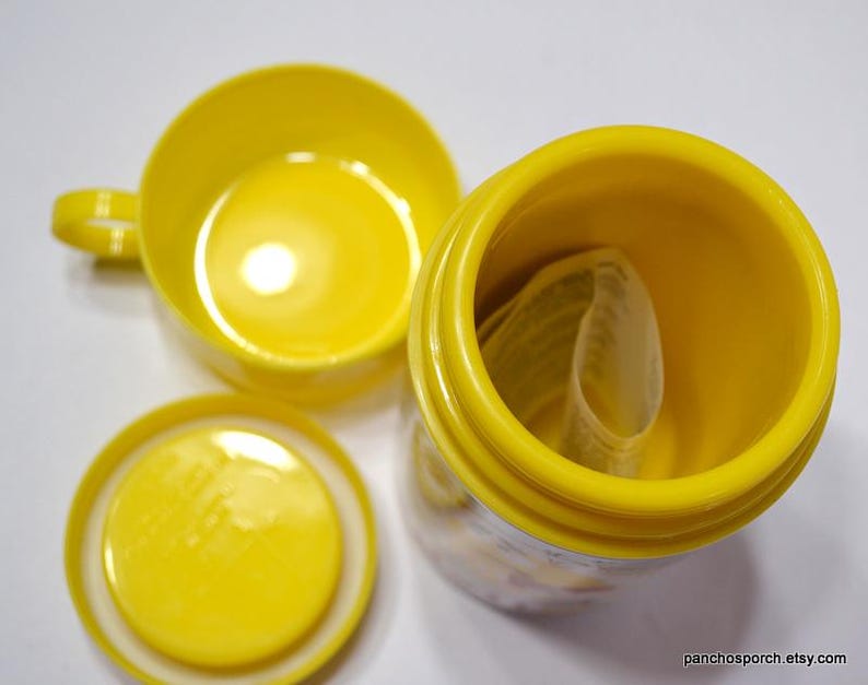 May include: Bright yellow ceramic cup with a handle, a matching lid, and a cylindrical container. The container is open, revealing a rolled-up paper inside. The items are arranged on a white surface.