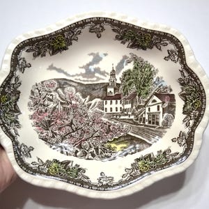 May include: An oval ceramic bowl with a scalloped edge featuring a cream-colored base. The center displays a detailed transfer print of a village scene with a church, trees, and pink blossoms. A brown and green floral border frames the scene.
