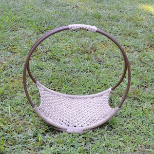 May include: A circular brown metal frame featuring a beige macrame woven base and a matching beige wrapped handle. The structure is designed with a curved, open-top basket shape, showcasing a detailed knotted cord pattern throughout the center.