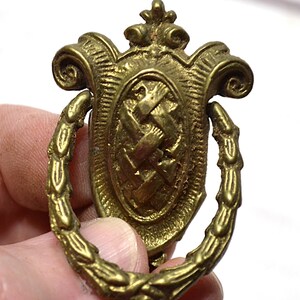 Peut inclure: Une poignée de tiroir en métal doré avec une plaque ovale ornée d'un motif tressé central. La quincaillerie comprend une poignée tombante décorative articulée en forme de couronne de laurier, finie avec des volutes ornementales au sommet.