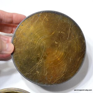 Op de afbeelding: Een rond metalen object in messingkleur met een donkere rand. Het oppervlak heeft een cirkelvormig patroon en talrijke krassen. Het object wordt door een hand vastgehouden, wat de grootte aangeeft. De algehele uitstraling is vintage.