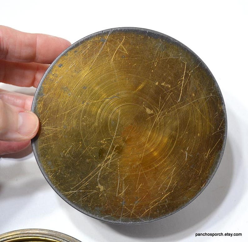 Op de afbeelding: Een rond metalen object in messingkleur met een donkere rand. Het oppervlak heeft een cirkelvormig patroon en talrijke krassen. Het object wordt door een hand vastgehouden, wat de grootte aangeeft. De algehele uitstraling is vintage.