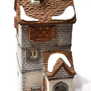 May include: A detailed ceramic miniature house with a brown tiled roof and a red brick chimney. The house has multiple levels with faux brick siding, wooden shutters, and a small arched entryway. White snow accents the roof.