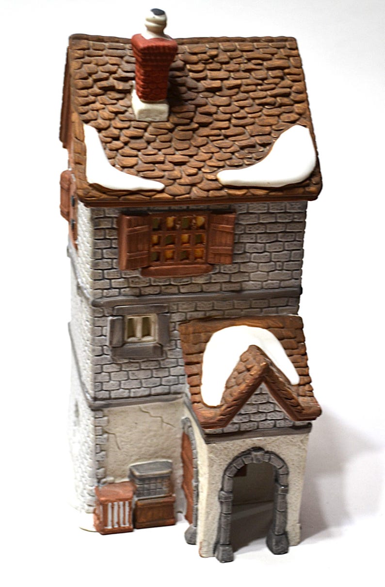 May include: A detailed ceramic miniature house with a brown tiled roof and a red brick chimney. The house has multiple levels with faux brick siding, wooden shutters, and a small arched entryway. White snow accents the roof.