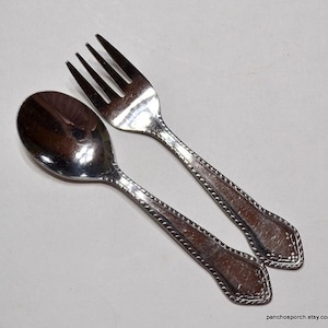 May include: A silver spoon and fork set. The spoon has a rounded bowl, and the fork has four tines. Both utensils have decorative patterned handles. The set is made of shiny metal and is placed on a light-colored surface.