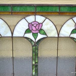 Op de afbeelding: Een glas-in-loodraam met drie gebogen panelen met roze rozen met groene bladeren. De panelen worden gescheiden door dunne zwarte metalen frames. De achtergrond is een wit, ondoorzichtig glas met een getextureerd oppervlak.