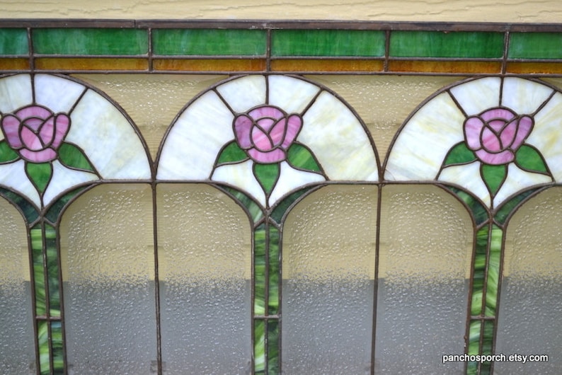 Op de afbeelding: Een glas-in-loodraam met drie gebogen panelen met roze rozen met groene bladeren. De panelen worden gescheiden door dunne zwarte metalen frames. De achtergrond is een wit, ondoorzichtig glas met een getextureerd oppervlak.