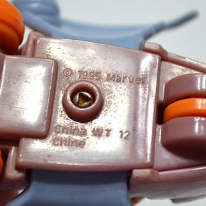 May include: Close-up of a section of a toy, featuring a mauve-colored plastic base with orange and gray accents. The text "&copy; 1995 Marvel" and "China WT 12 Chine" are visible. The toy appears to be a vehicle or a part of a larger playset.