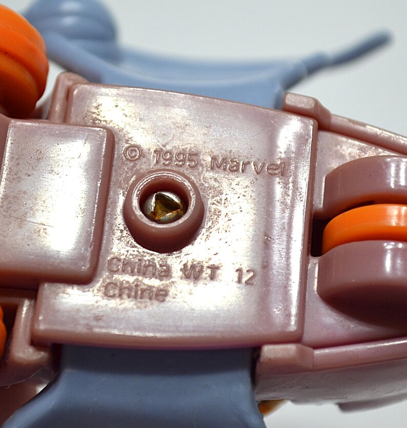 May include: Close-up of a section of a toy, featuring a mauve-colored plastic base with orange and gray accents. The text "&copy; 1995 Marvel" and "China WT 12 Chine" are visible. The toy appears to be a vehicle or a part of a larger playset.