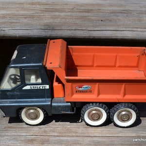 May include: A vintage Structo toy dump truck featuring a dark gray cab and a bright orange hydraulic dump bed. The truck has white-walled wheels with silver hubcaps, a white interior with a steering wheel, and the word Structo printed on the side.