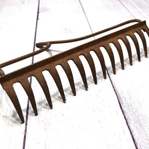 May include: An antique, rusty metal garden rake with a long handle and multiple tines. The rake is made of iron and has a curved handle for easy grip. The tines are pointed and designed for raking leaves and debris.