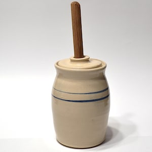 May include: A cream-colored ceramic crock with a matching lid and two horizontal blue stripes around the center. A cylindrical wooden handle protrudes vertically from the center of the lid, extending upward from the top of the vessel.