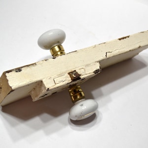 May include: A rectangular, off-white painted wooden block featuring two smooth, white porcelain door knobs attached with brass-colored metal hardware. The paint on the wooden block shows signs of wear, cracking, and chipping throughout.
