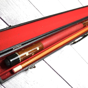 May include: A vintage pool cue set in a black case with a red velvet interior. The cue is brown with white dot inlays and a red grip. A second cue is also visible. The case is open, revealing the cues.