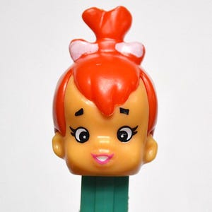 May include: A close-up of a Fred Flintstone character PEZ dispenser. The dispenser features a cartoon head with orange hair, a pink bow, and a green base. The character has large eyes and a pink smile.