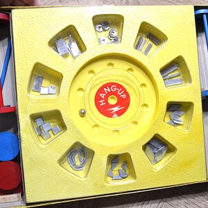 May include: A vintage Hang-Up board game in a yellow plastic tray with a central red circular label featuring the text HANG-UP and a lightning bolt. The tray contains various metal game pieces, red and blue plastic rods, and circular discs.