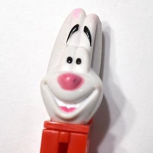 May include: A white plastic Pez candy dispenser featuring the head of a cartoon rabbit with long ears, black eyebrows, black eyes, and a pink nose and mouth. The dispenser body is a solid orange-red rectangular plastic piece.