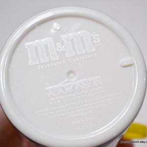 May include: White insulated container with the embossed text "m&m's" and "Insulated Container". The bottom of the container also features the text "A. ARONSON" and other text. The container is held by a hand.