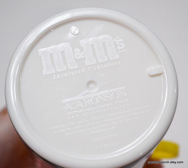May include: White insulated container with the embossed text "m&m's" and "Insulated Container". The bottom of the container also features the text "A. ARONSON" and other text. The container is held by a hand.