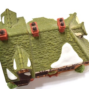 May include: A miniature decorative house with a green roof, red chimneys, and white snow accents. The house has a detailed, textured roof and is likely part of a holiday village display.