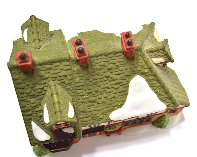 May include: A miniature decorative house with a green roof, red chimneys, and white snow accents. The house has a detailed, textured roof and is likely part of a holiday village display.