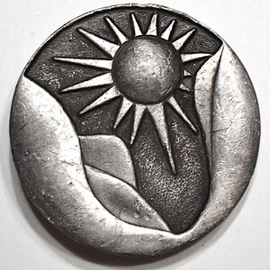 May include: A round, silver-toned metal button or pendant featuring a sunburst design with a central sun and radiating rays. The sun is set against a backdrop of stylized mountain shapes, creating a nature-inspired theme.