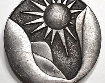 Könnte beinhalten: Ein runder, silberfarbener Metallknopf oder Anhänger mit einem Sonnenstrahl-Design mit einer zentralen Sonne und strahlenden Strahlen. Die Sonne ist vor einem Hintergrund aus stilisierten Bergformen platziert, was ein von der Natur inspiriertes Thema schafft.